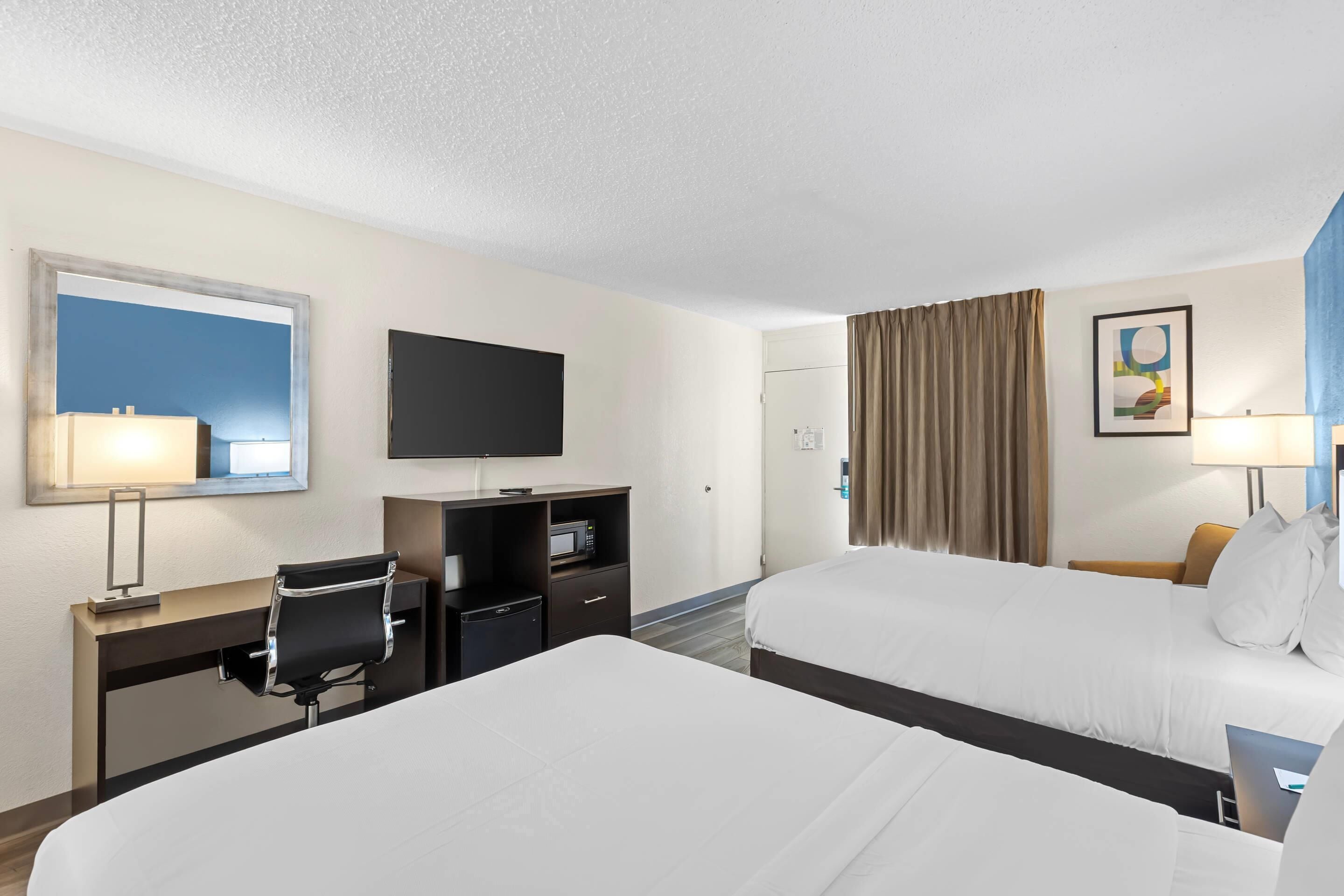 A hotel room with two beds, a desk with a chair, a flat-screen TV, a desk lamp, a microwave, and a window with curtains.