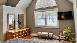 A bedroom with a sloped ceiling, a large window with blinds, a wooden dresser, a bench, a television mounted on the wall, and a potted plant. - Southern Vermont Retreat w/ Hot Tub, Fire Pit, & Bunk Room (Windham)