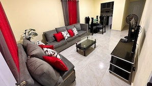 Een moderne woonkamer met een grijze bank, rode en witte kussens, een glazen salontafel en een tv-meubel met een flatscreen televisie.