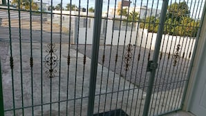 Een metalen hekwerk met decoratieve details, dat leidt naar een verhard gebied met een witte schutting en gebouwen op de achtergrond.