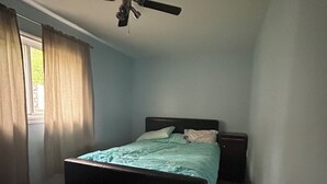 Un dormitorio con cama, ventilador de techo, ventana con cortinas y piso de madera.