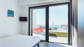 Ein Schlafzimmer mit einem Bett, einem Fernseher und einer Glastür, die auf einen Balkon mit Blick auf das Meer und ein Gebäude führt.