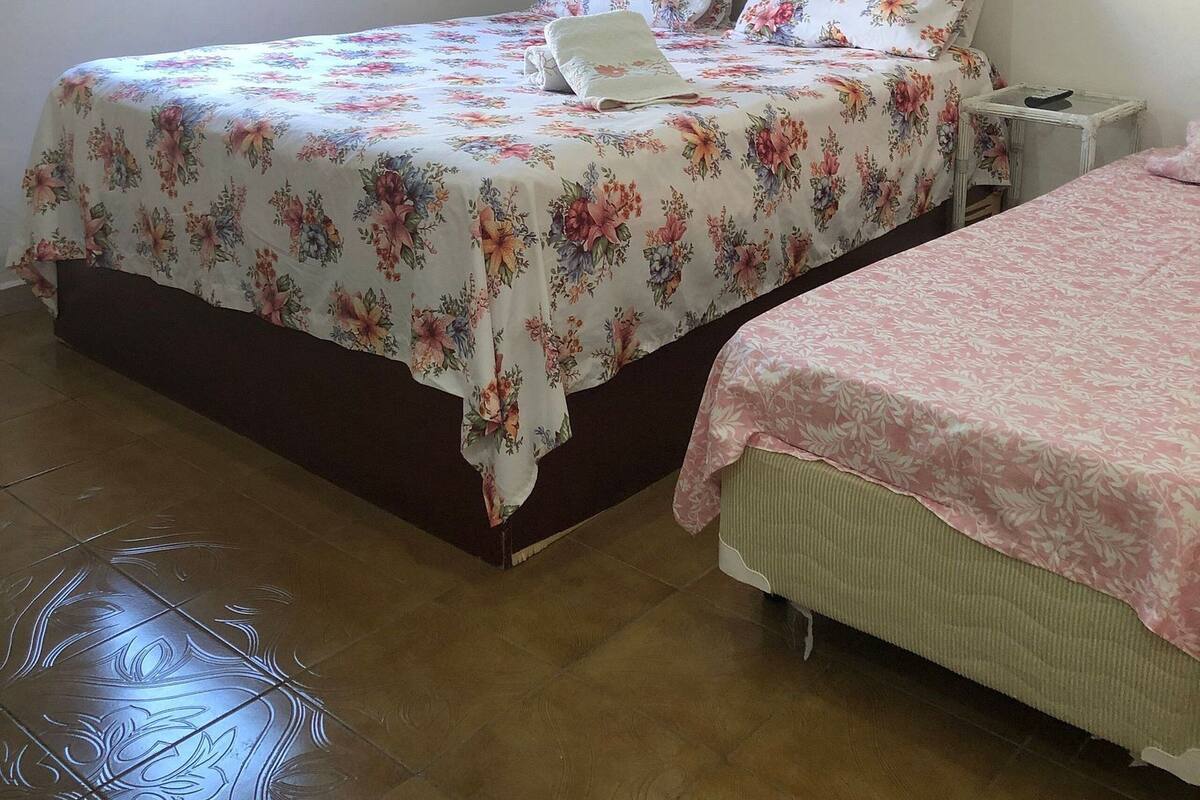 Egy hálószoba virágmintás ágytakaróval, egy éjjeliszekrény egy lámpával, és egy ablak, amelyen redőny van.