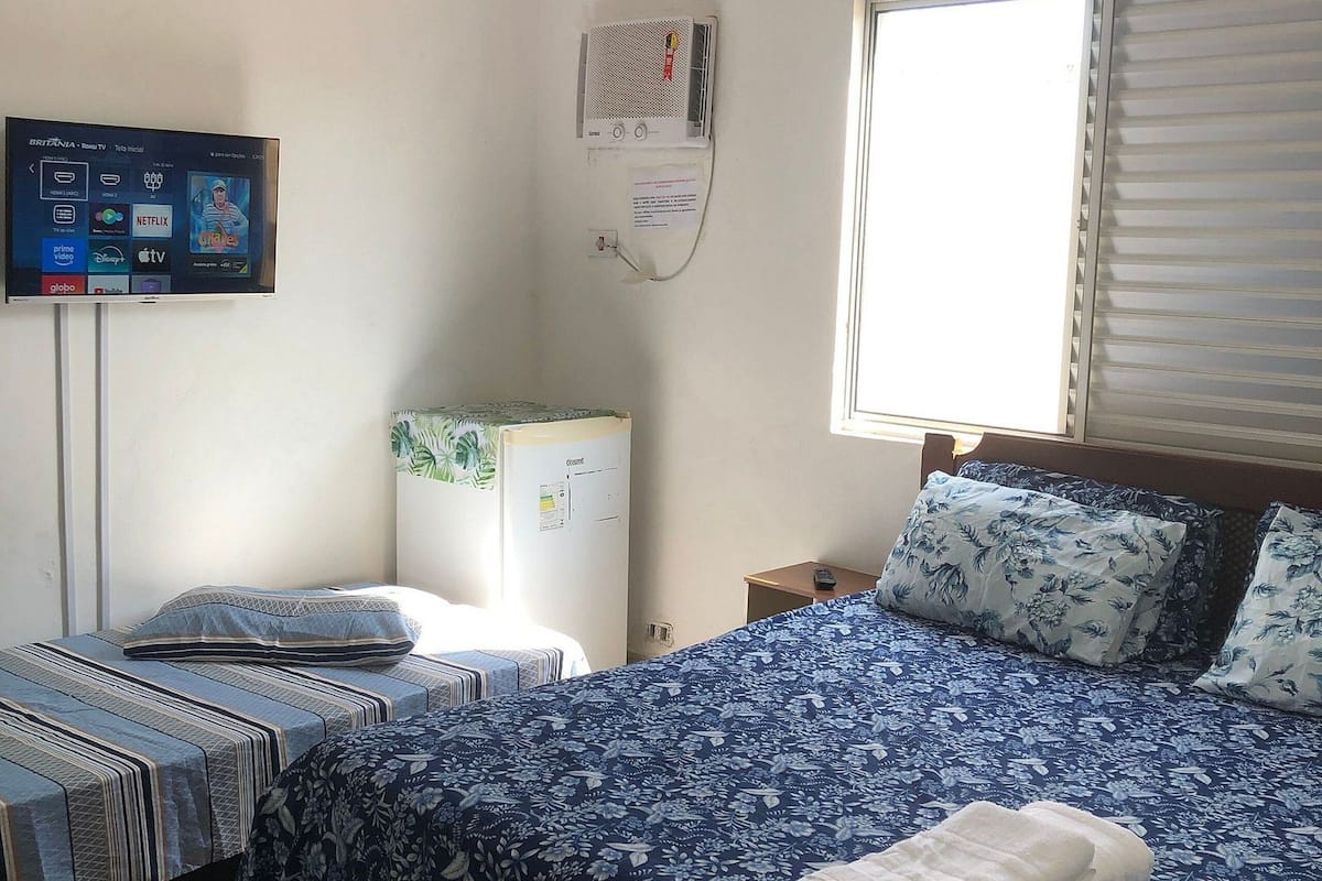 Egy szállodai szoba, amelyben található egy ágy, egy kis asztal, egy falra szerelt légkondicionáló, egy televízió és egy ablak, amelyen zsaluzi van.
