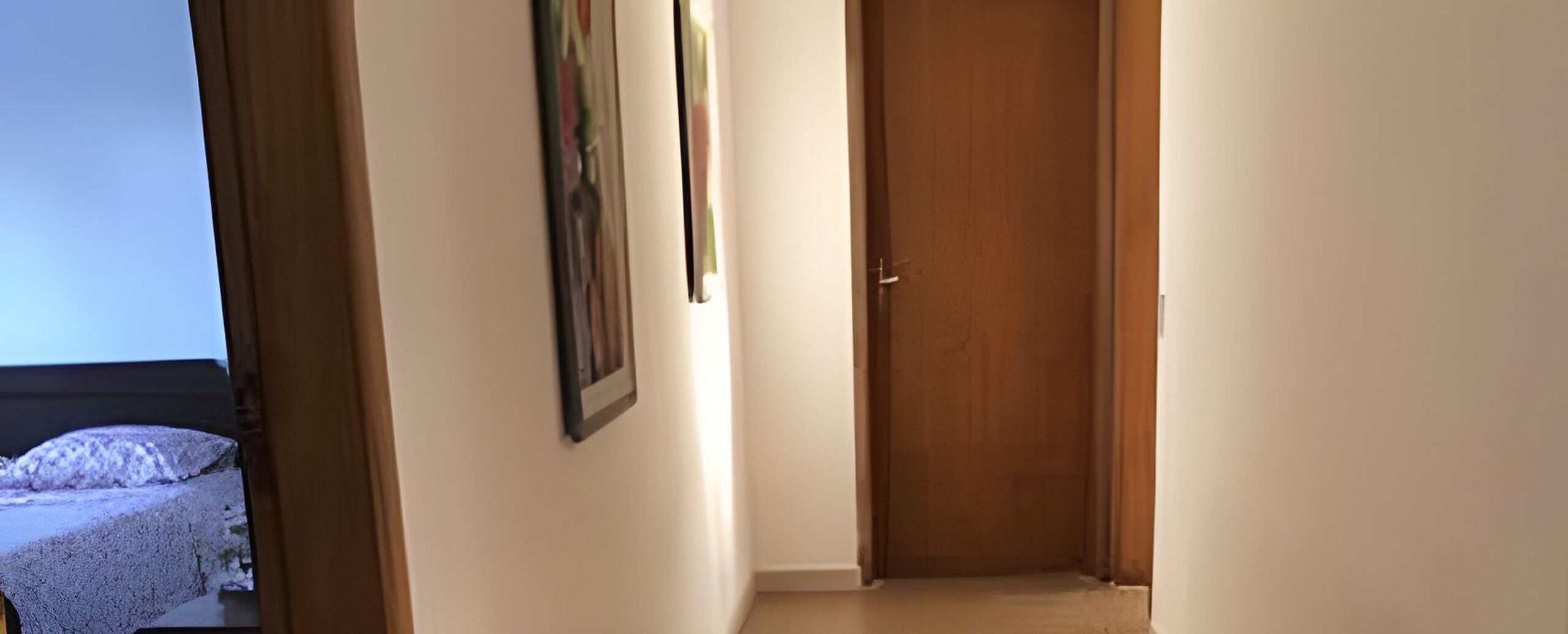 Um corredor com uma porta de madeira, um quadro na parede e uma cama com cobertura branca ao fundo.