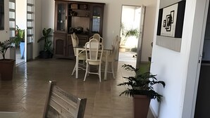 Ένας ευρύχωρος χώρος καθιστικού με τραπέζι και καρέκλες για το φαγητό, μια κουζίνα με ντουλάπια και γλάστρες με φυτά.