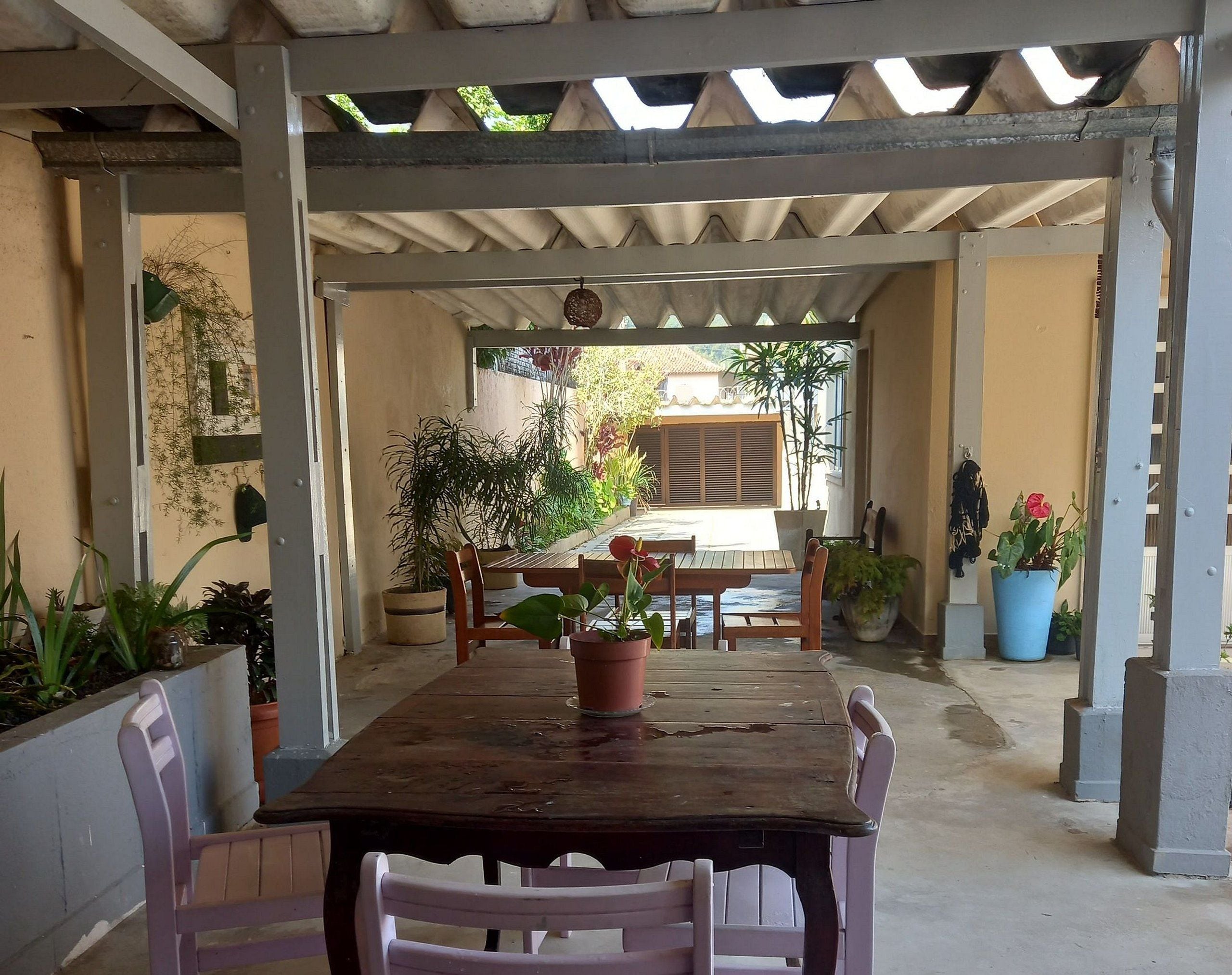 A covered outdoor dining area with a wooden table and chairs, surrounded by potted plants.