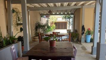 A covered outdoor dining area with a wooden table and chairs, surrounded by potted plants.