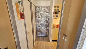 Un pasillo estrecho con una puerta decorada con un papel tapiz tropical. Se observa un mueble pequeño con puertas de vidrio al fondo.