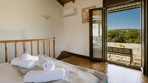 A bedroom with a bed, a wooden headboard, a balcony with a table and chairs, and a view of greenery and hills.