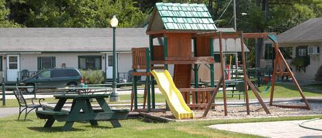 Un parque infantil con tobogán, columpios, mesas de picnic y una pequeña construcción.