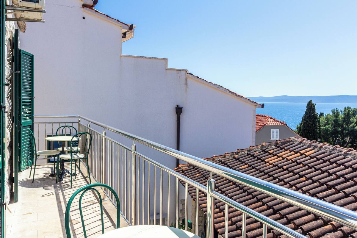 A balcony with a table and chairs, a metal railing, and a view of the sea and mountains.