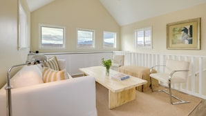 Un salon moderne avec un plafond haut, un ventilateur de plafond, une grande fenêtre, un canapé blanc, une table basse en bois et un tableau encadré accroché au mur.