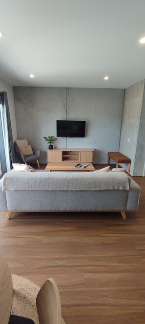 Uma sala de estar moderna com um sofá cinza grande, uma mesa de centro de madeira, uma TV de tela plana na parede, uma pequena mesa lateral de madeira e uma poltrona cinza.