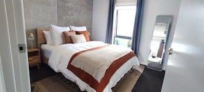Quarto de hotel moderno com uma cama grande, uma mesa de cabeceira de madeira, um abajur e vista para o exterior através de uma janela.