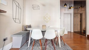 A dining area with a white table, four white chairs, and a grey bench. There are abstract wall art pieces and a door leading to another room. - 3bedroom Design Gem in Kensington (London)