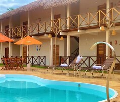 Uma área de piscina com espreguiçadeiras e guarda-sóis, piscina e um edifício com telhado de palha e varandas de madeira.