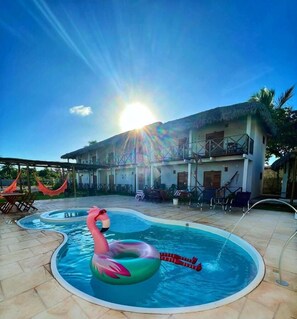 En swimmingpool med en uppblåsbar flamingofigur, en byggnad med halmtak och utomhusmöbler.