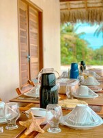 Mesa lista con platos, vasos y una cafetera, con una decoración de estrella de mar y vistas al exterior con vegetación.