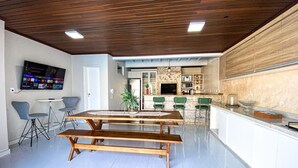 A modern kitchen with a wooden countertop, a dining area with a wooden table and benches, and a flat-screen TV mounted on the wall. - Casa com piscina 5 suites 1 quarto e AC (Foz do Iguaçu)