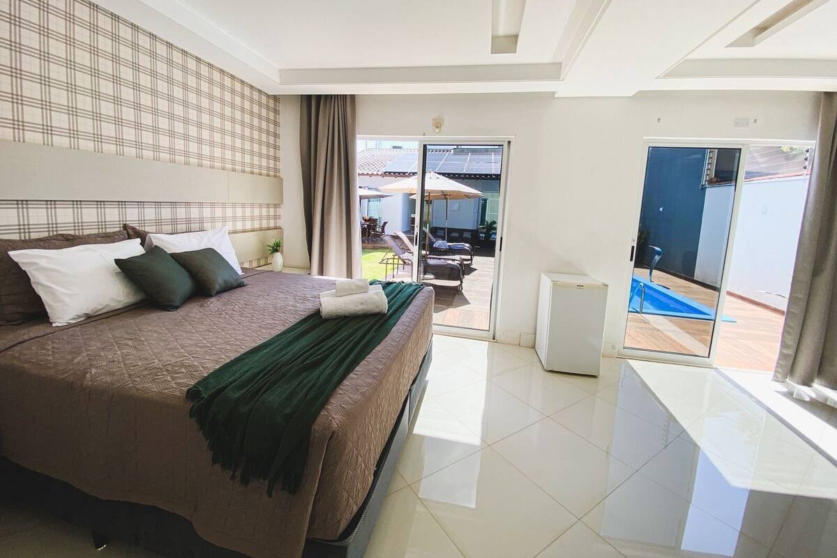 Ein Hotelzimmer mit einem großen Bett, einem kleinen Schreibtisch, einer Glastür zu einer Terrasse mit Sitzgelegenheiten und einer Deckenleuchte.