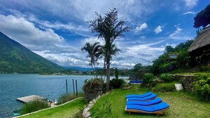 Vista a un lago con un muro de piedra, una palmera y sillones azules para relajarse.