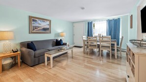 Un salon comprenant un canapé, une table basse et une table à manger avec des chaises. Il y a un tableau au mur, une lampe sur une table d’appoint et un panier sous le canapé.