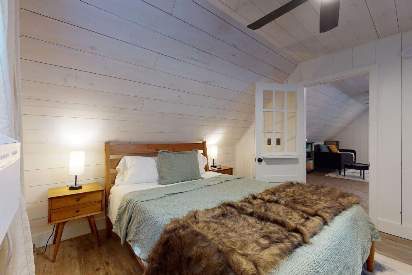 Un dormitorio con cama de madera, lámparas de noche, un cubrecama de piel y vista a un salón comedor a través de una puerta abierta.