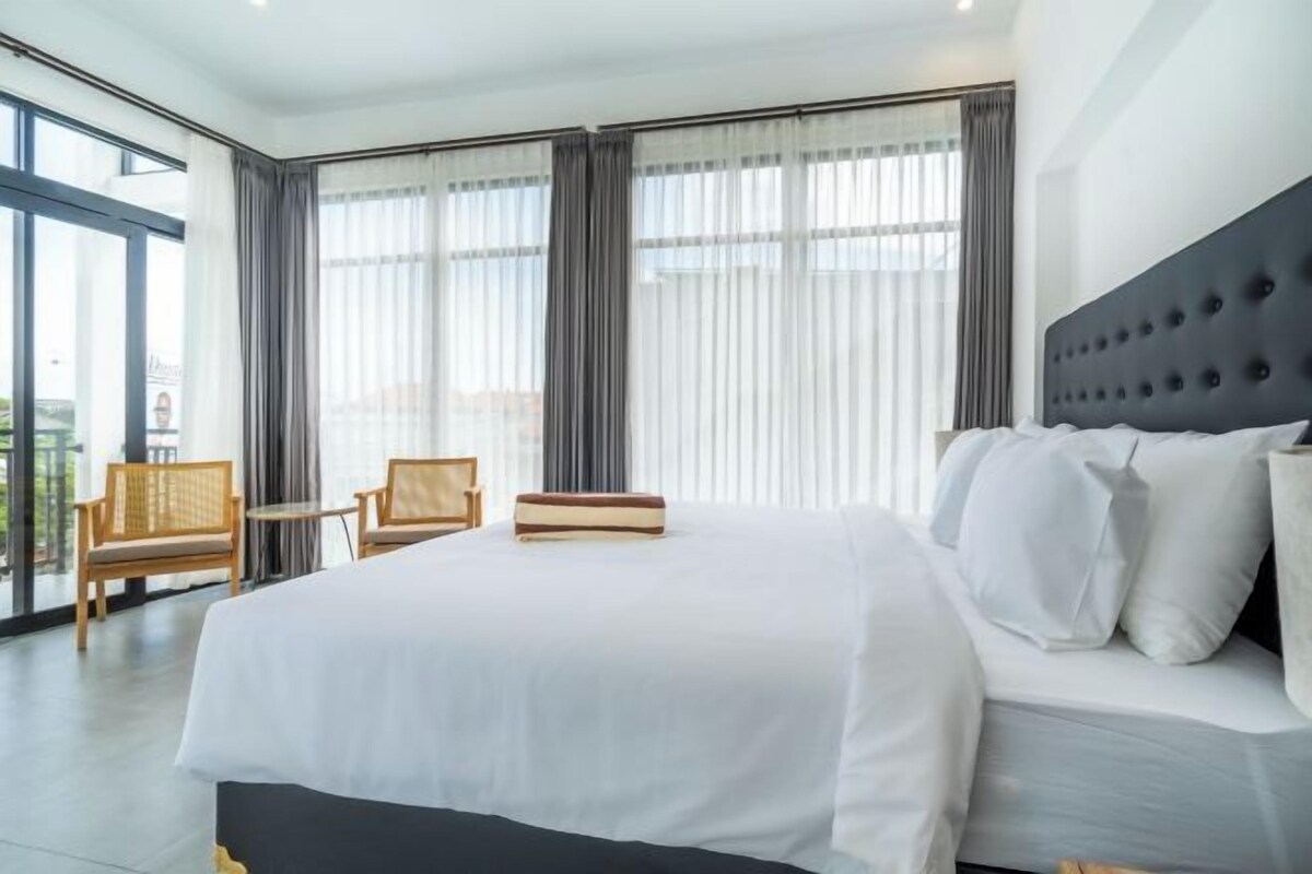 A modern hotel room with a large bed, a small table, two chairs, and a view of the city through large windows.