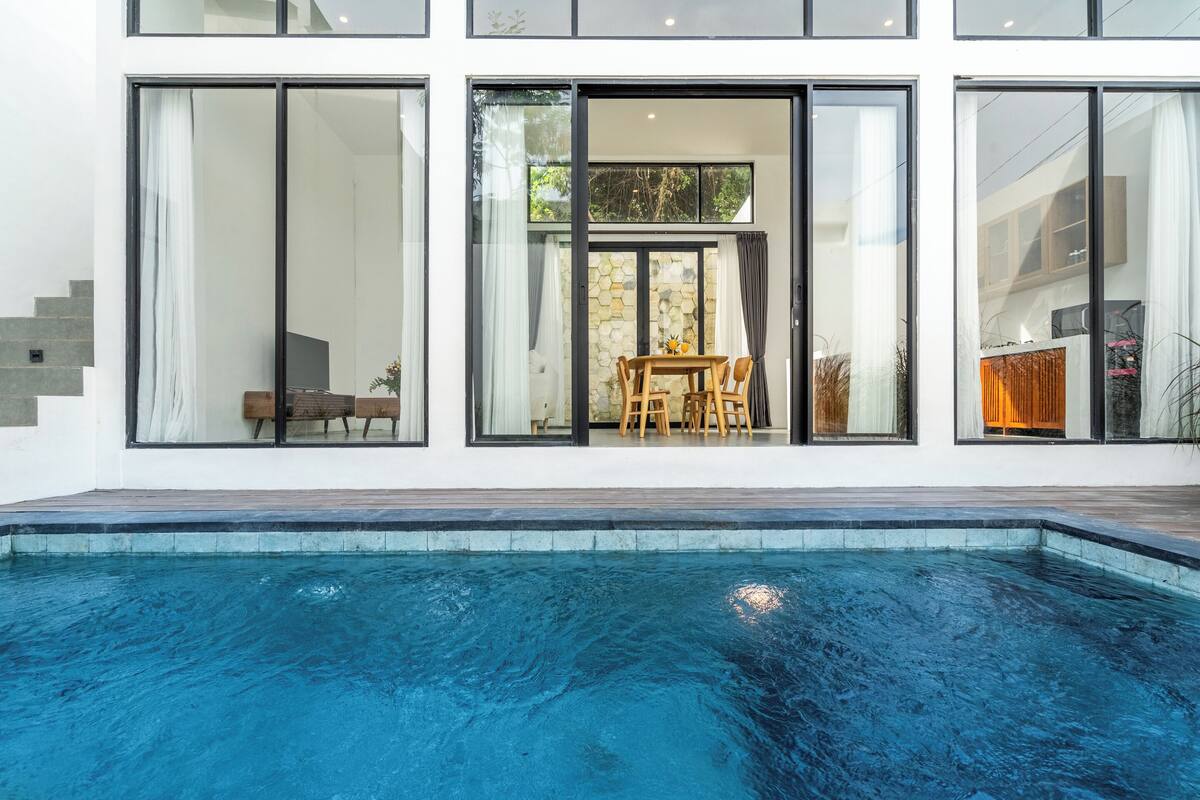 A modern house with a swimming pool, large windows, and a dining area visible through the glass doors.