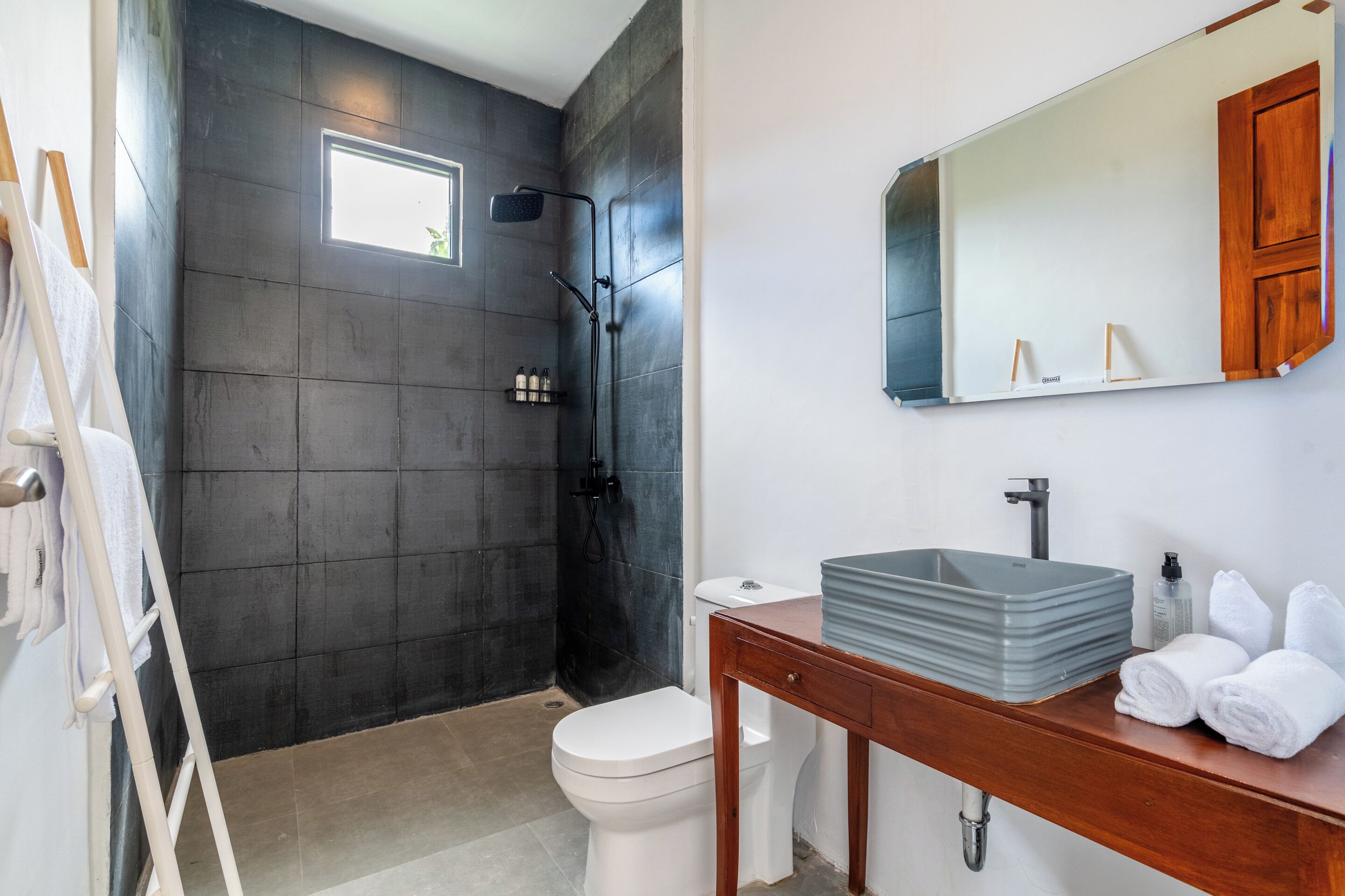 A bathroom with a dark tiled wall, a white toilet, a shower with a black hose, a wooden vanity with a sink, and a mirror.