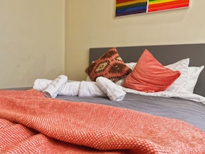 Een bed met een grijze matras, witte kussens en een rood deken met een patroon.