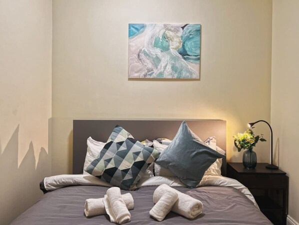 Ein ordentlich bezogenes Bett mit Kissen, einer Nachttischlampe und einem an der Wand hängenden abstrakten Gemälde.