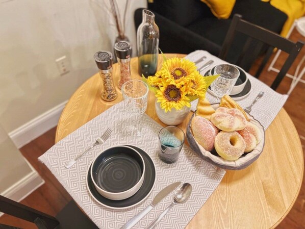 Une table de salle à manger ronde, recouverte d’une nappe à motifs, un bol rempli de beignets, un vase avec des tournesols, des salières et des poivrières, et un verre d’eau.