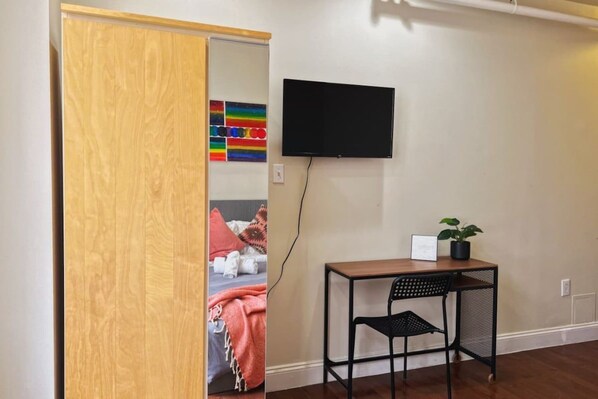 A room with a wooden door, a bed with a colorful blanket, a desk with a chair, a potted plant, and a mounted television.