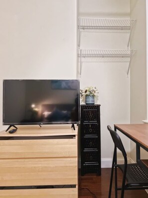 Un televisor sobre un soporte de madera, un altavoz negro, una mesa de comedor con una silla y una pared blanca con una repisa metálica.