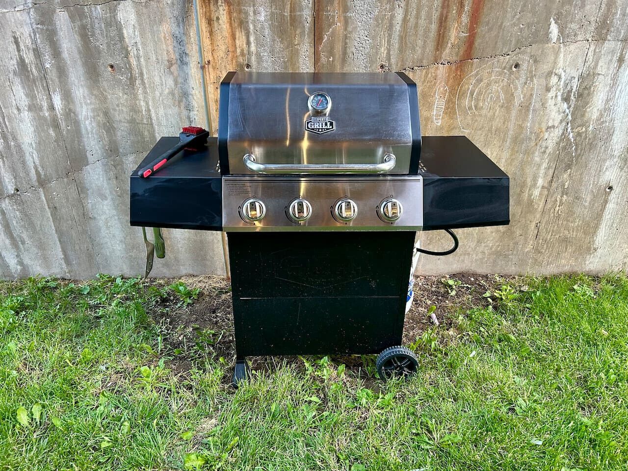 Un barbecue au gaz portatif, doté de quatre brûleurs et d’une étagère latérale, est placé sur l’herbe, adossé à une clôture en bois.