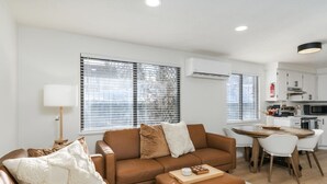 Een moderne woonkamer met een bruine leren bank, een houten salontafel en een eethoek met witte stoelen.