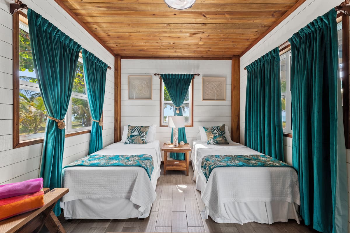 Ein Zimmer mit zwei Betten, türkisblauen Vorhängen, einer Holzbalkendecke und einem kleinen Tisch mit Lampe.
