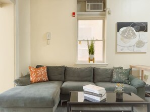 Una sala de estar con un sofá gris, una mesa de centro con libros, una ventana con una planta y un cuadro en la pared.
