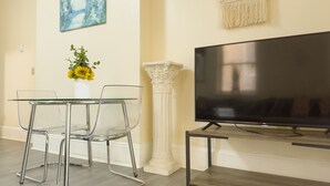 Una sala de estar moderna con una mesa de vidrio, dos sillas transparentes, un televisor sobre un soporte de madera, un jarrón con girasoles, un cuadro y un adorno de pared.