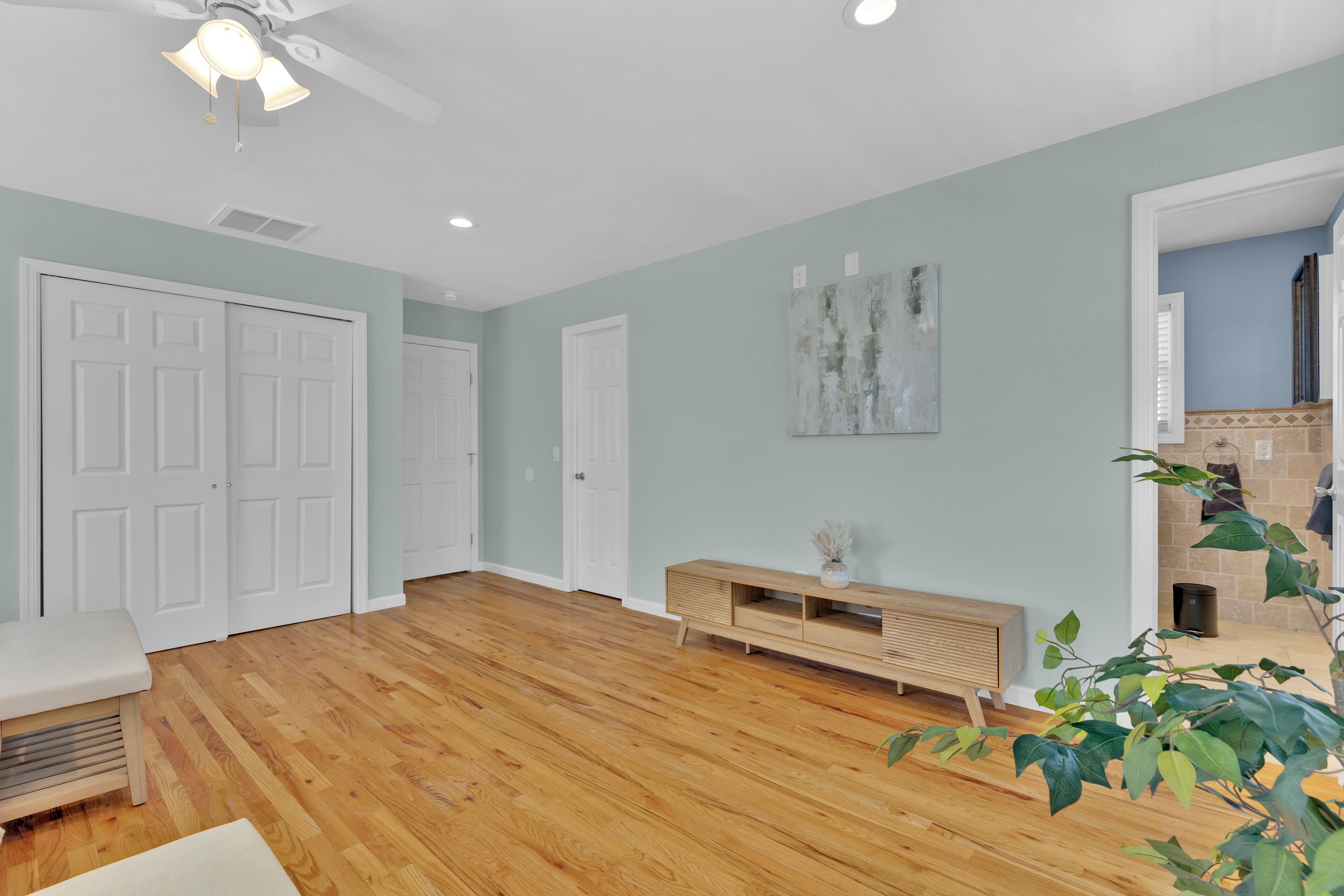 A spacious living room with hardwood flooring, white doors, a wooden bench, a console table, and a ceiling fan.