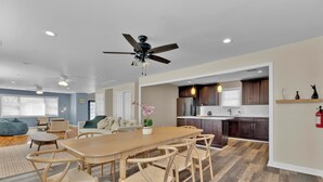 A spacious living room with a large wooden dining table and matching chairs, a kitchen with dark cabinets, and a ceiling fan.