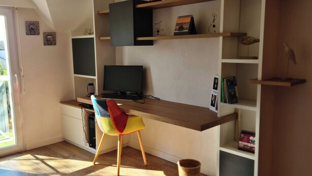 Una oficina en casa moderna con escritorio, computadora y silla de colores. Se ven estantes con objetos decorativos y una planta pequeña.