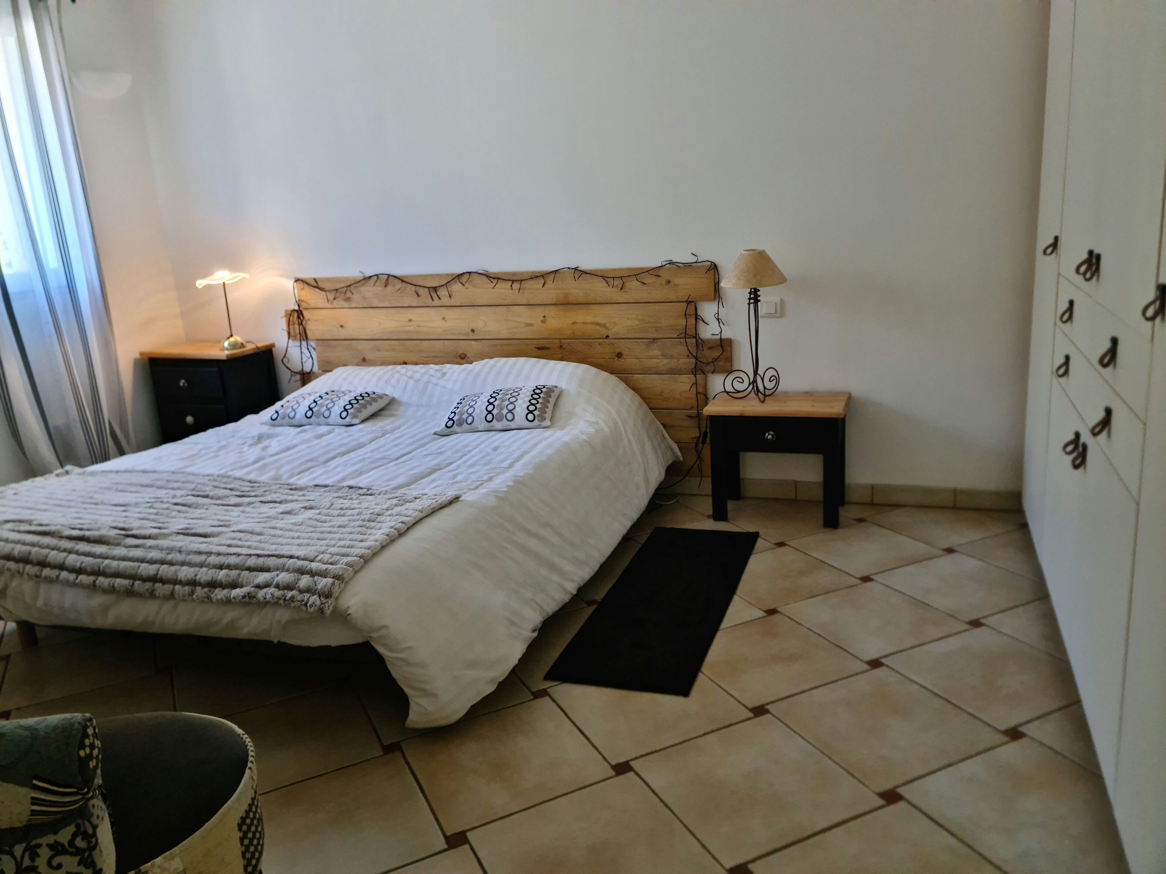 A neatly arranged bedroom with a wooden headboard, a bed with patterned pillows, a nightstand with a lamp, and a small table with a lamp.