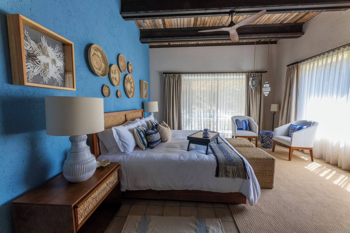 Un dormitorio con una cama grande, una mesita de noche con lámpara, una silla y un ventanal con cortinas.