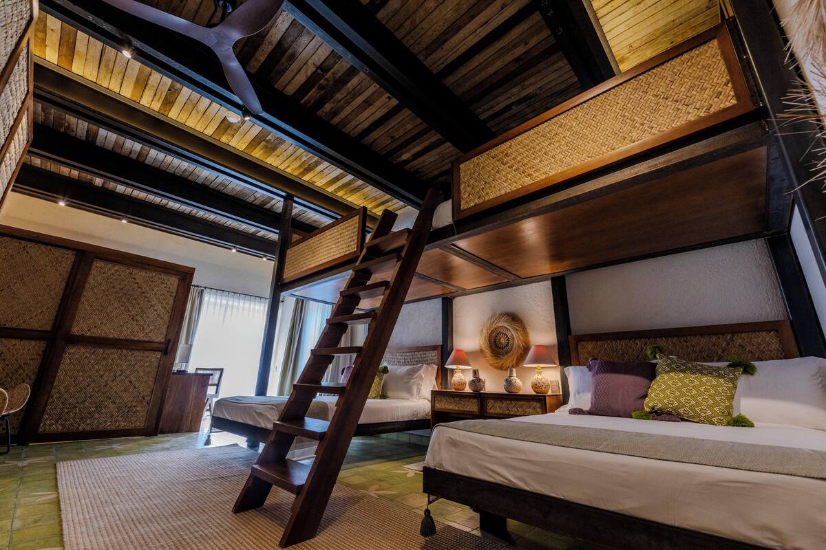 Una habitación con techo de madera, una cama con ropa blanca, una mesita de noche con lámparas y una escalera que conduce a un nivel superior.