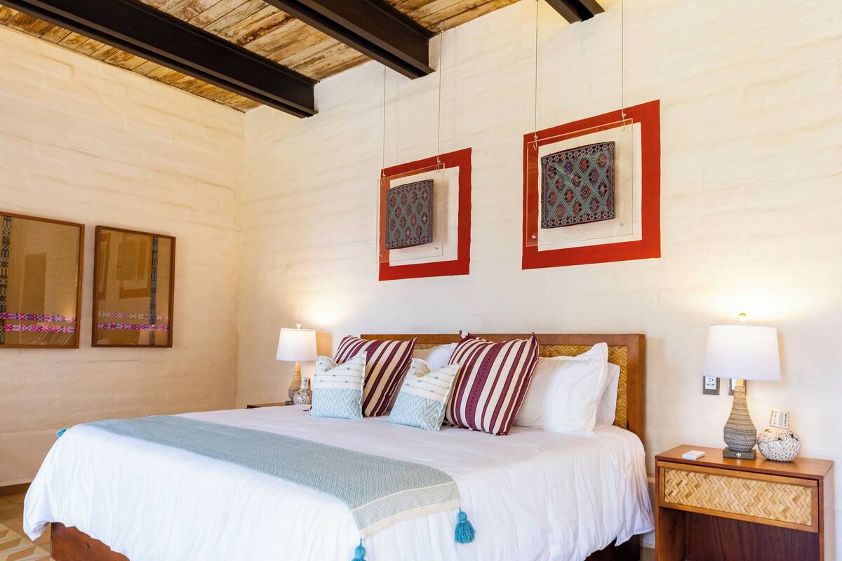 Un dormitorio con techo de madera, una cama con ropa de cama blanca, dos mesitas de noche con lámparas y cuadros de pared con diseño.