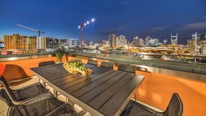 Una terraza en la azotea con una mesa y sillas, con vistas a un horizonte urbano nocturno.