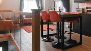 A modern living room with a glass railing, a dining area with two orange chairs, a flat-screen TV, and a kitchen area with a copper-colored cabinet. - 1 bedroom accommodation in Windermere (Windermere)